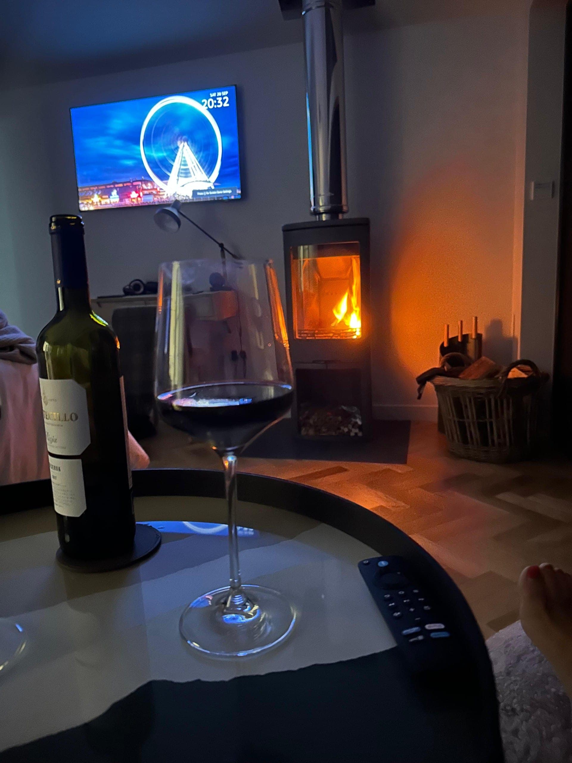 A glass of red wine by the lit wood-burning stove, with a film on the TV